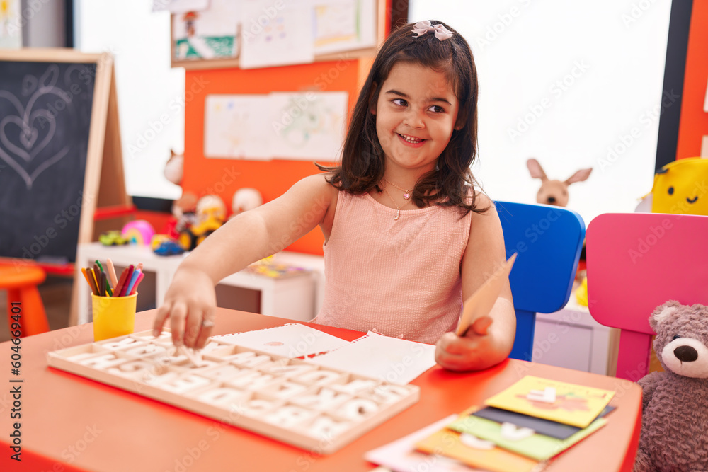 Fototapeta premium Adorable hispanic girl playing with vocabulary puzzle game sitting on table at kindergarten