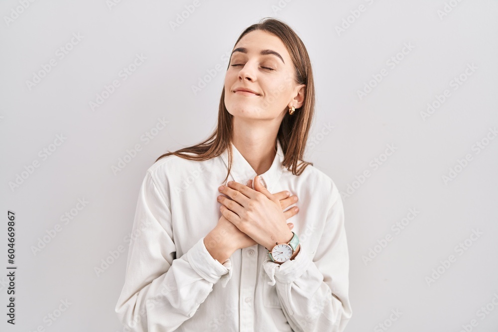 Young caucasian woman standing over isolated background smiling with hands on chest with closed eyes and grateful gesture on face. health concept. Young caucasian woman standing over isolated background smiling with hands on chest with closed eyes and grateful gesture on face. health concept.
