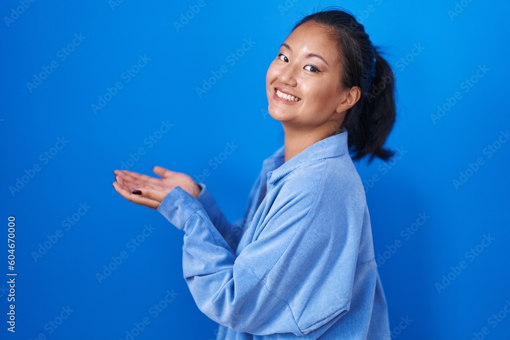Asian young woman standing over blue background pointing aside with hands open palms showing copy space, presenting advertisement smiling excited happy Asian young woman standing over blue background pointing aside with hands open palms showing copy space, presenting advertisement smiling excited happy