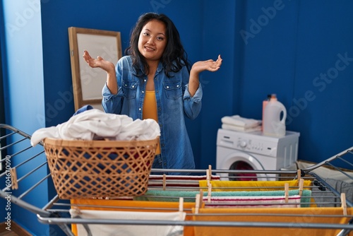 Young asian woman hanging clothes at clothesline clueless and confused expression with arms and hands raised. doubt concept.