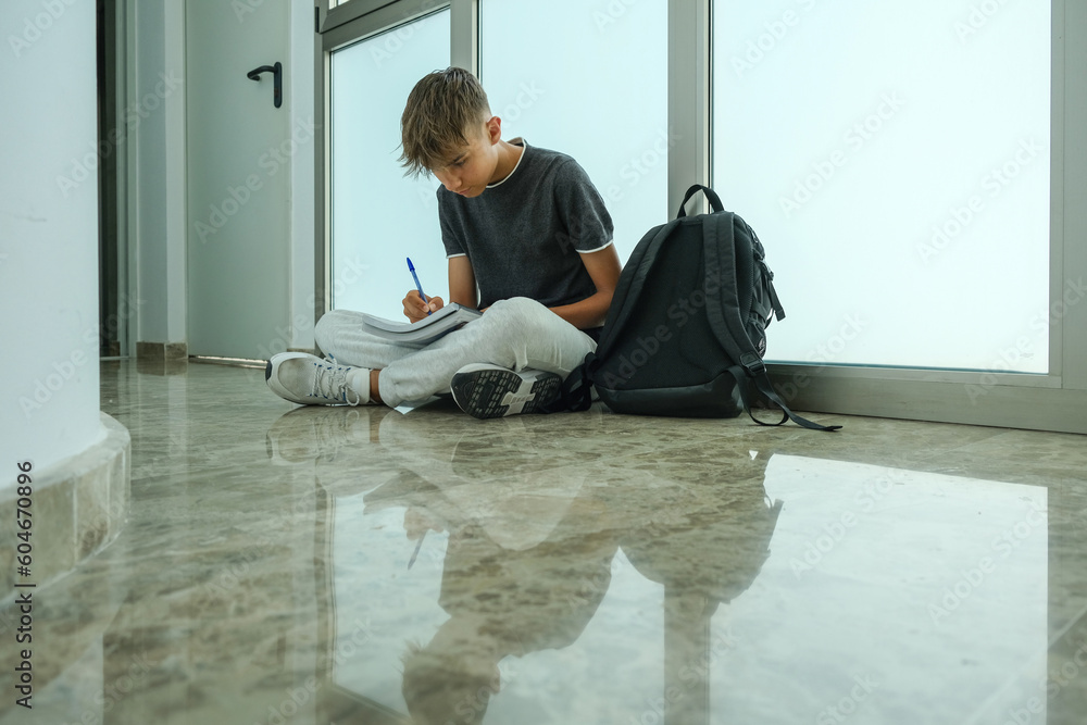 Teenage boy student wearing casual clothes sitting on floor and writing, makes notes in notebook. Teenager studies, learning, writing, doing homework, preparing to exam