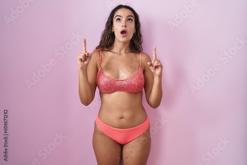 Young hispanic woman wearing lingerie over pink background amazed and surprised looking up and pointing with fingers and raised arms.
