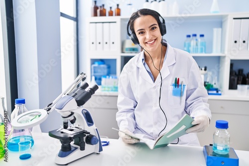 Young hispanic woman wearing scientist uniform listening to music reading book at laboratory