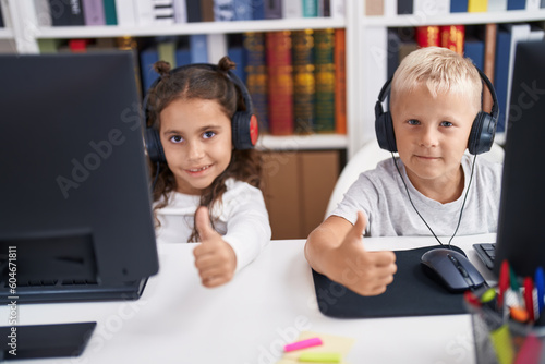 Adorable boy and girl students using computer and headphones doing thumbs up gesture at classroom