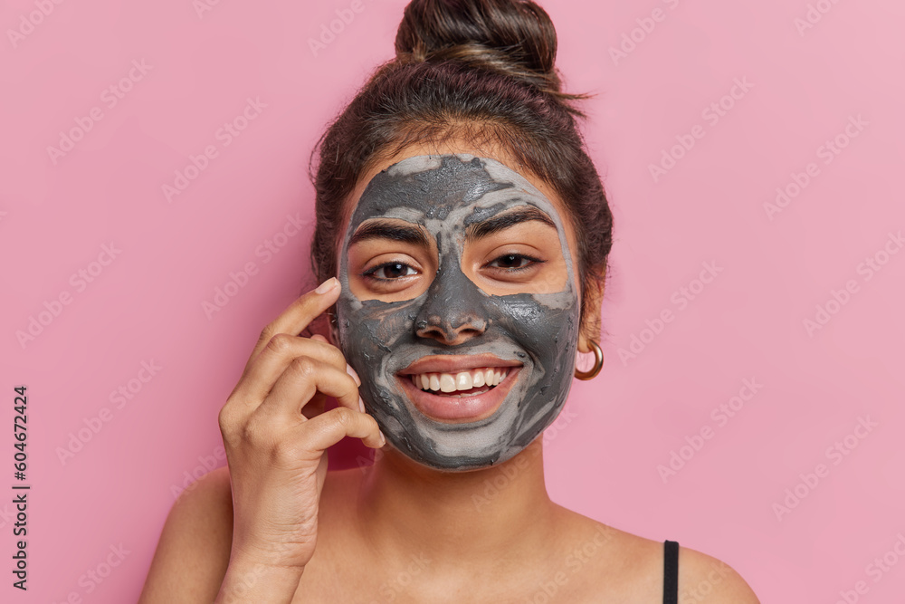 Portrait of good looking cheerful woman applies cosmetic clay mask smiles toothily looks happily at camera stands bare shoulders isolated over pink background enjoys softness. Cosmetology treatment