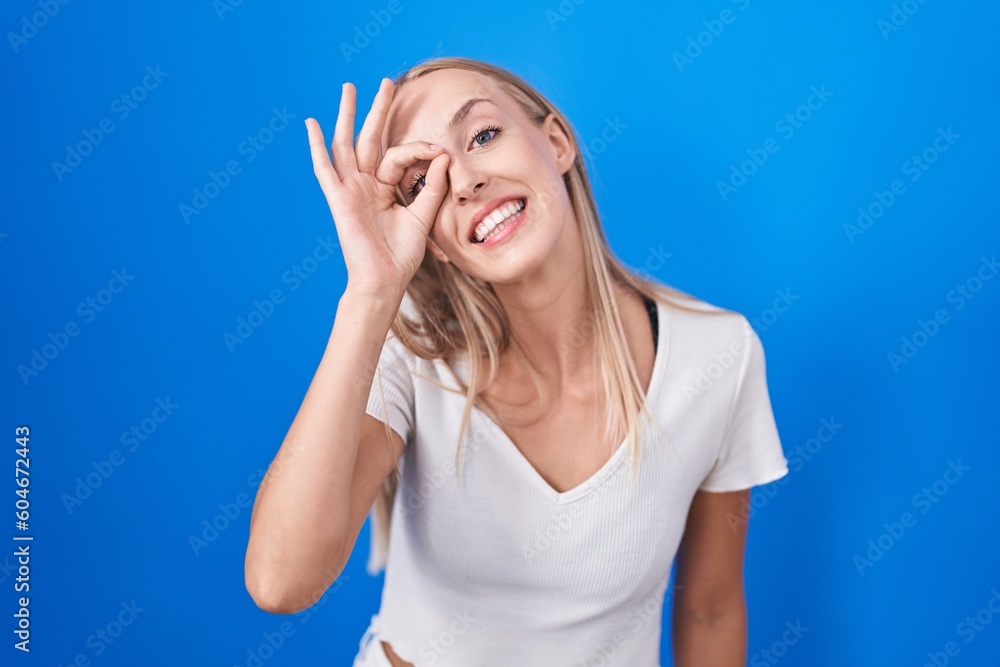 Young caucasian woman standing over blue background doing ok gesture with hand smiling, eye looking through fingers with happy face. Young caucasian woman standing over blue background doing ok gesture with hand smiling, eye looking through fingers with happy face.