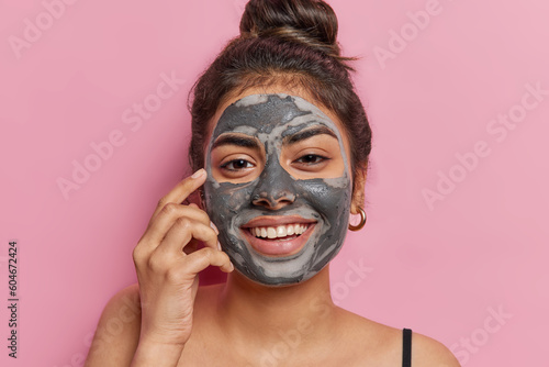 Portrait of good looking cheerful woman applies cosmetic clay mask smiles toothily looks happily at camera stands bare shoulders isolated over pink background enjoys softness. Cosmetology treatment