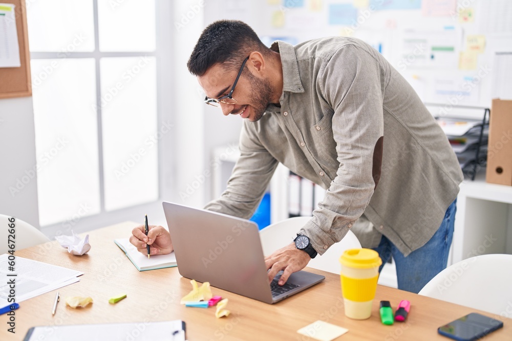 Young hispanic man business worker using laptop writing on notebook at office Young hispanic man business worker using laptop writing on notebook at office