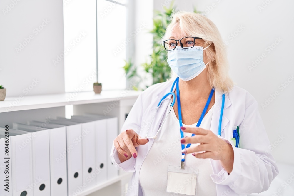 Middle age blonde woman wearing doctor uniform and medical mask speaking at clinic Middle age blonde woman wearing doctor uniform and medical mask speaking at clinic
