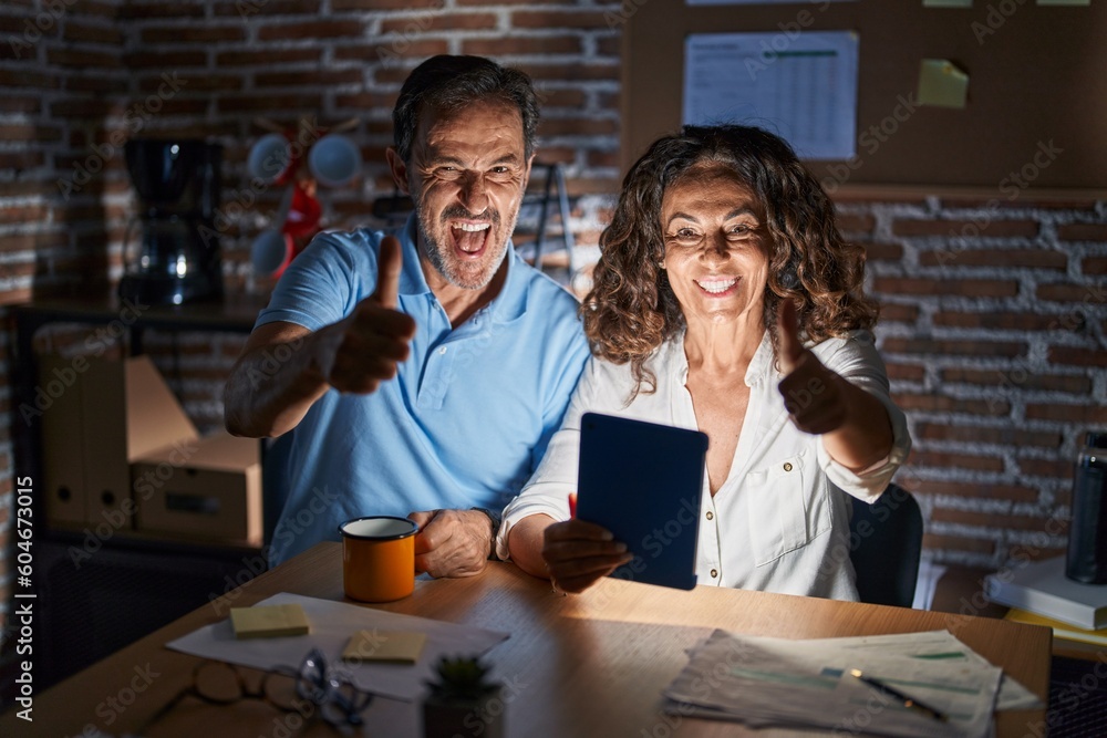 Middle age hispanic couple using touchpad sitting on the table at night approving doing positive gesture with hand, thumbs up smiling and happy for success. winner gesture. Middle age hispanic couple using touchpad sitting on the table at night approving doing positive gesture with hand, thumbs up smiling and happy for success. winner gesture.