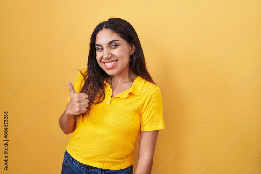 Young arab woman standing over yellow background doing happy thumbs up gesture with hand. approving expression looking at the camera showing success. Young arab woman standing over yellow background doing happy thumbs up gesture with hand. approving expression looking at the camera showing success.