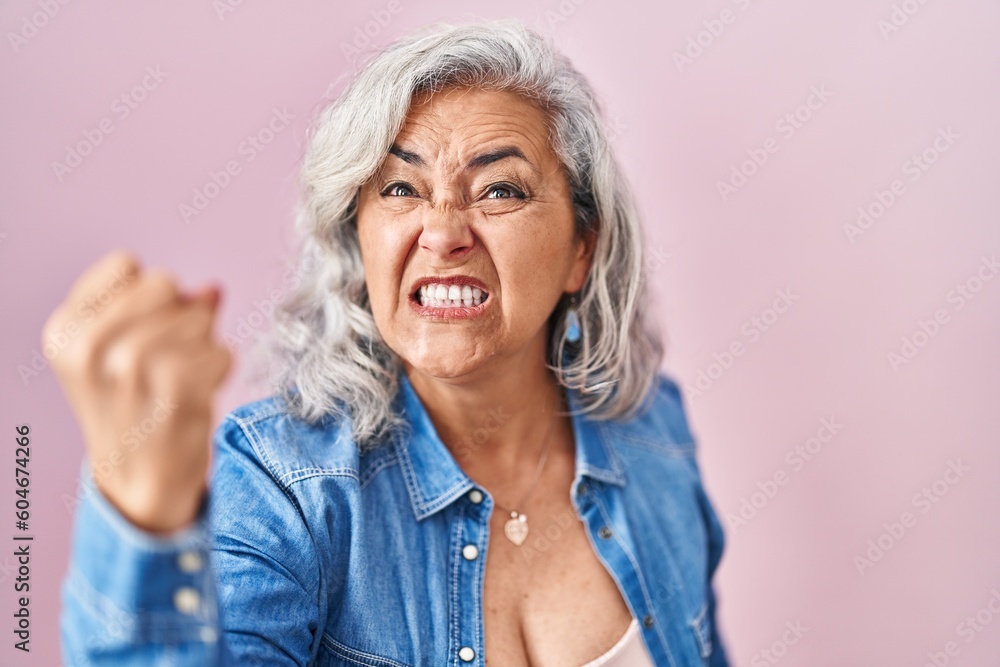 Middle age woman with grey hair standing over pink background angry and mad raising fist frustrated and furious while shouting with anger. rage and aggressive concept. Middle age woman with grey hair standing over pink background angry and mad raising fist frustrated and furious while shouting with anger. rage and aggressive concept.