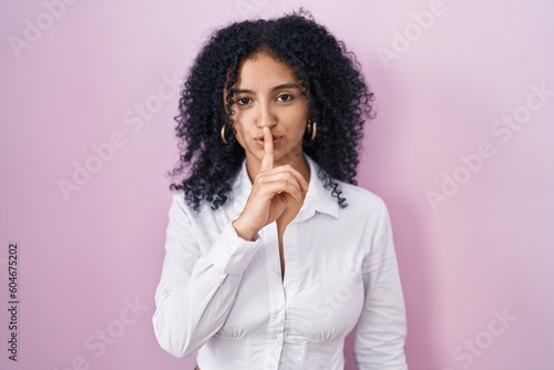 Hispanic woman with curly hair standing over pink background asking to be quiet with finger on lips. silence and secret concept.
