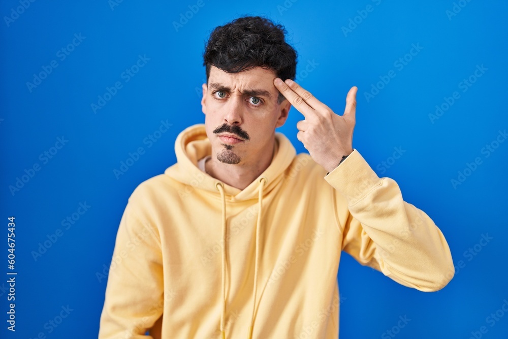 Hispanic man standing over blue background shooting and killing oneself pointing hand and fingers to head like gun, suicide gesture. Hispanic man standing over blue background shooting and killing oneself pointing hand and fingers to head like gun, suicide gesture.