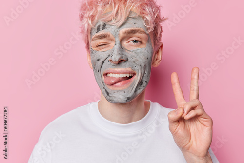 Funny man winks eye sticks out tongue shows peace sign foolishes around applies beauty clay mask for reducing acnes dressed in casual white t shirt isolated over pink background. Skin care concept