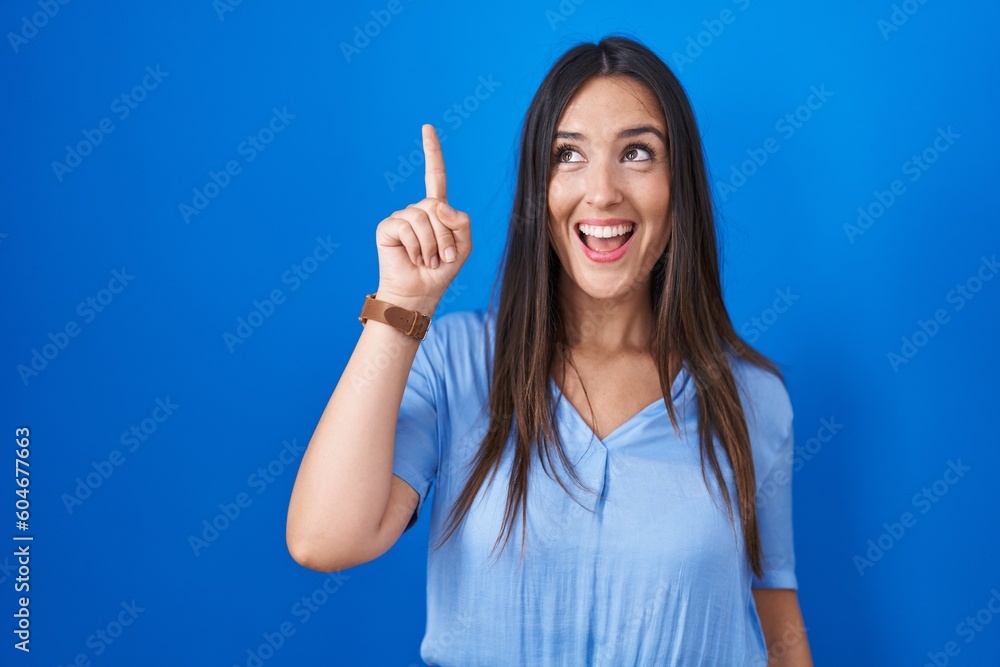 Young brunette woman standing over blue background pointing finger up with successful idea. exited and happy. number one. Young brunette woman standing over blue background pointing finger up with successful idea. exited and happy. number one.