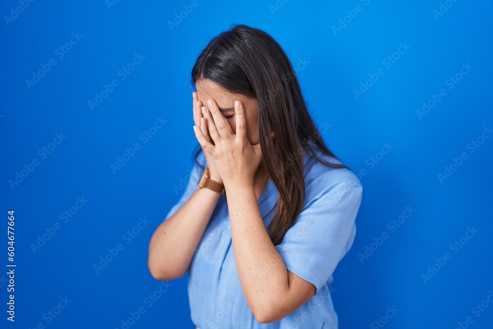 Young brunette woman standing over blue background with sad expression covering face with hands while crying. depression concept. Young brunette woman standing over blue background with sad expression covering face with hands while crying. depression concept.