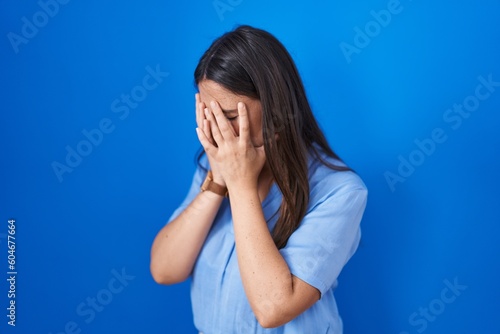 Young brunette woman standing over blue background with sad expression covering face with hands while crying. depression concept.