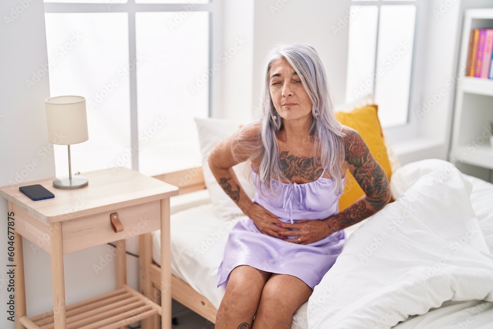 Middle age grey-haired woman suffering for stomach ache sitting on bed at bedroom Middle age grey-haired woman suffering for stomach ache sitting on bed at bedroom