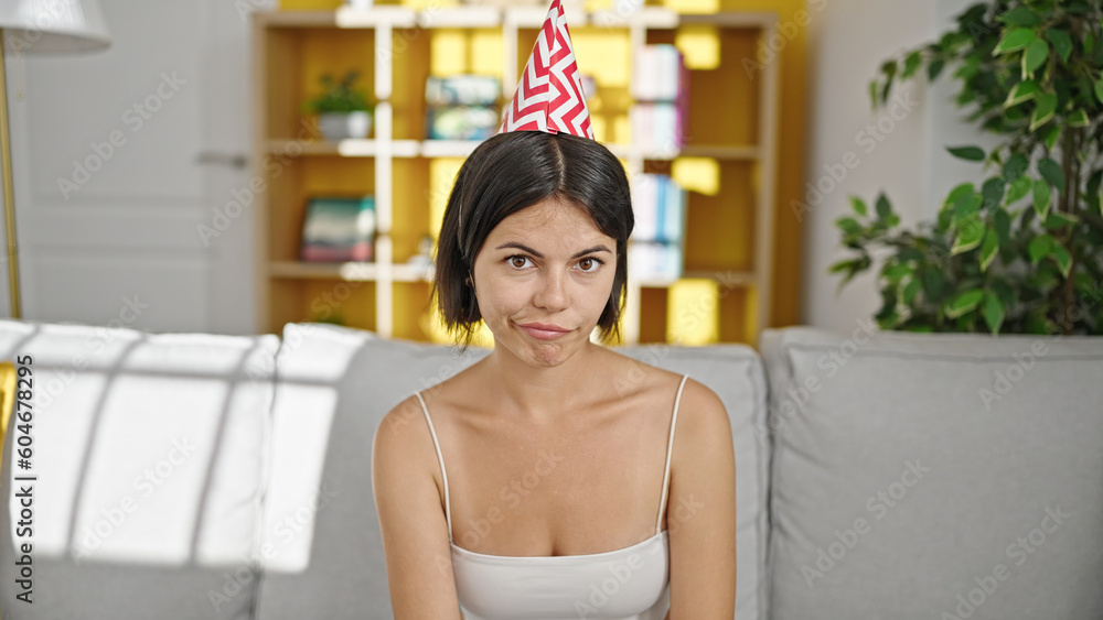 Young beautiful hispanic woman wearing birthday hat sitting on sofa at home Young beautiful hispanic woman wearing birthday hat sitting on sofa at home