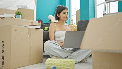 Young beautiful hispanic woman using laptop sitting on floor at new home