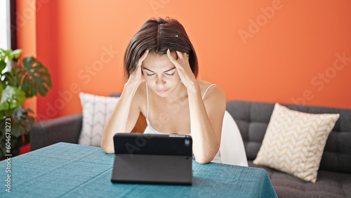 Young beautiful hispanic woman looking upset watching touchpad at dinning room