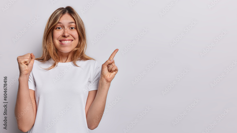 Horizontal shot of cheerful woman clenches fist and bites lips points index finger at copy space celebrates positive news dressed in casual t shirt isolated over white background. Wow great offer - obrazy, fototapety, plakaty 