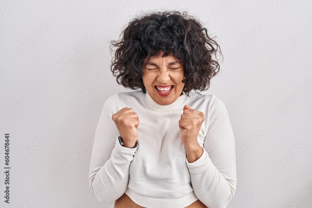 Hispanic woman with curly hair standing over isolated background excited for success with arms raised and eyes closed celebrating victory smiling. winner concept. Hispanic woman with curly hair standing over isolated background excited for success with arms raised and eyes closed celebrating victory smiling. winner concept.