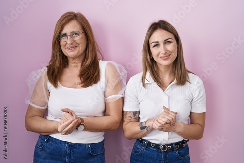 Hispanic mother and daughter wearing casual white t shirt over pink background with hands together and crossed fingers smiling relaxed and cheerful. success and optimistic