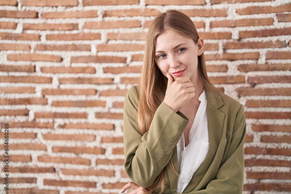 Young caucasian woman standing over bricks wall background looking confident at the camera smiling with crossed arms and hand raised on chin. thinking positive. Young caucasian woman standing over bricks wall background looking confident at the camera smiling with crossed arms and hand raised on chin. thinking positive.