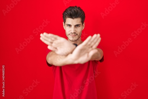 Young hispanic man standing over red background rejection expression crossing arms and palms doing negative sign, angry face