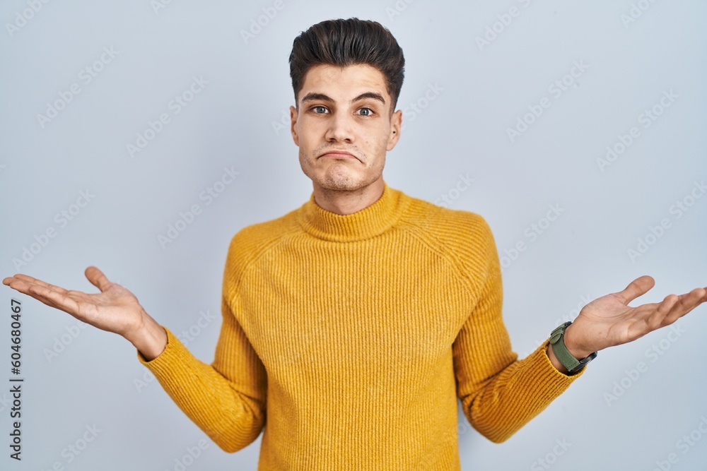 Young hispanic man standing over blue background clueless and confused expression with arms and hands raised. doubt concept. - obrazy, fototapety, plakaty 