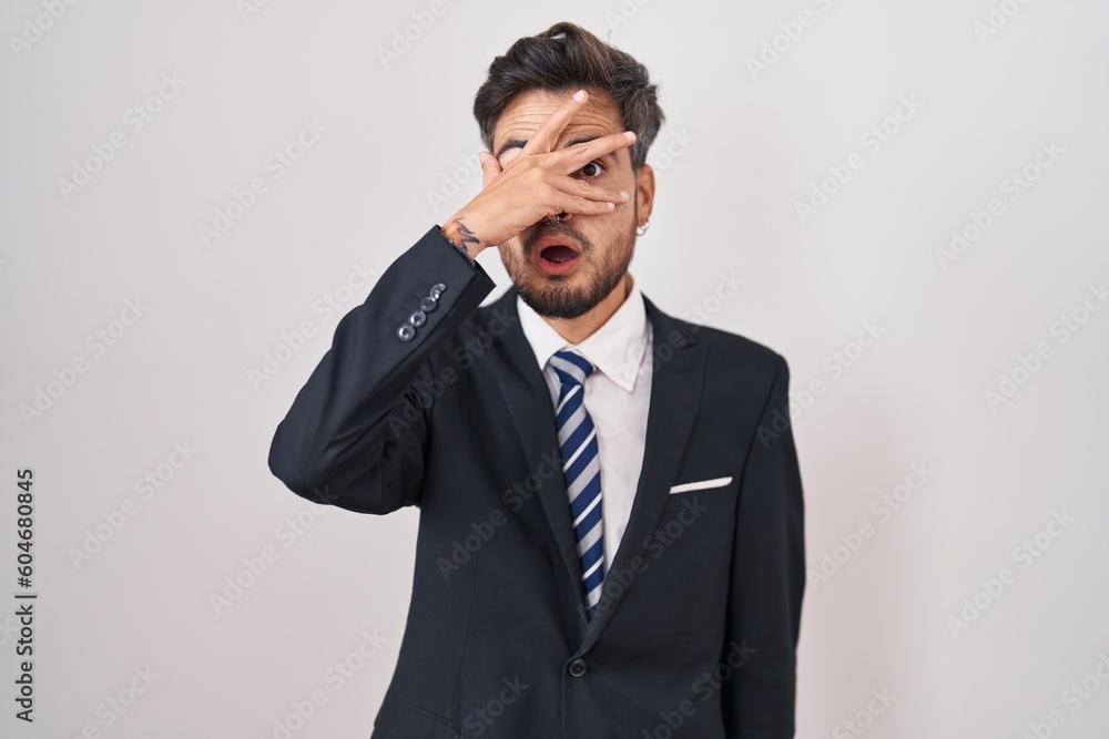Young hispanic man with tattoos wearing business suit and tie peeking in shock covering face and eyes with hand, looking through fingers with embarrassed expression. Young hispanic man with tattoos wearing business suit and tie peeking in shock covering face and eyes with hand, looking through fingers with embarrassed expression.