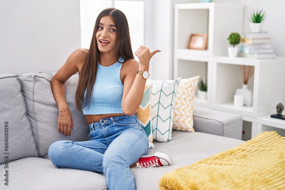 Young brunette woman sitting on the sofa at home smiling with happy face looking and pointing to the side with thumb up. Young brunette woman sitting on the sofa at home smiling with happy face looking and pointing to the side with thumb up.