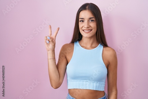 Young brunette woman standing over pink background showing and pointing up with fingers number two while smiling confident and happy.