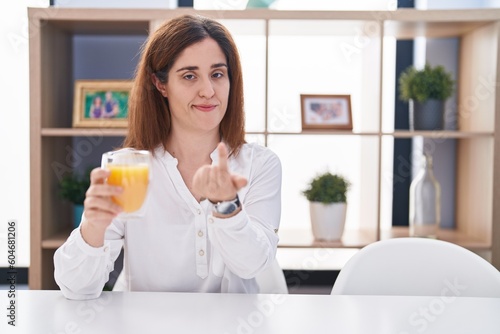 Brunette woman drinking glass of orange juice beckoning come here gesture with hand inviting welcoming happy and smiling