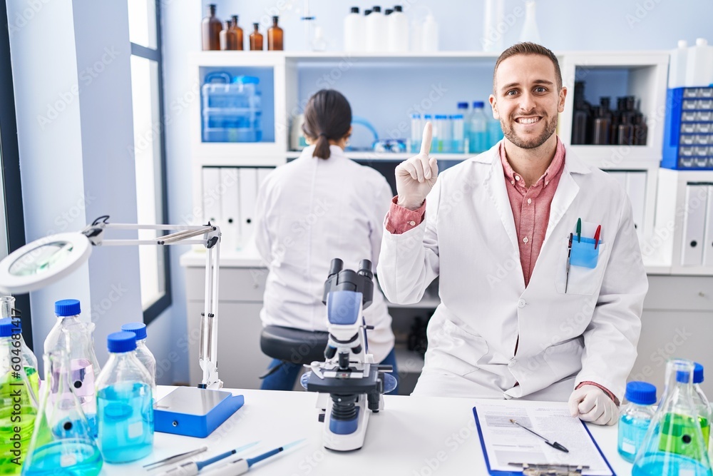 Young man working at scientist laboratory smiling with an idea or question pointing finger with happy face, number one Young man working at scientist laboratory smiling with an idea or question pointing finger with happy face, number one