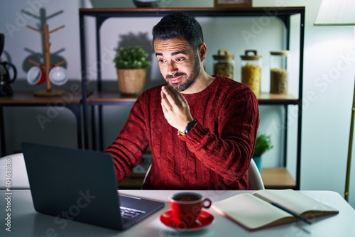 Young hispanic man with beard using computer laptop at night at home doing italian gesture with hand and fingers confident expression