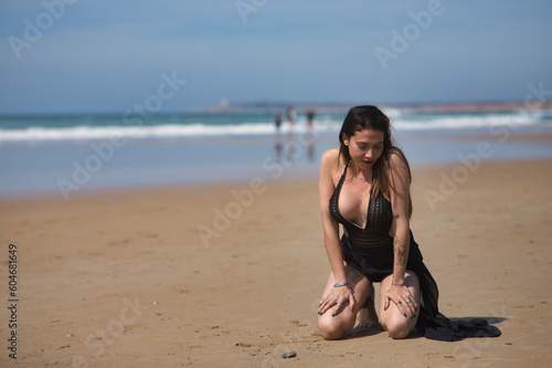 Attractive young woman  blue eyes  wearing black swimsuit and sarong  lonely  quiet and sad  kneeling on the shore of the beach. Concept tranquility  peace  sorrow  depression.