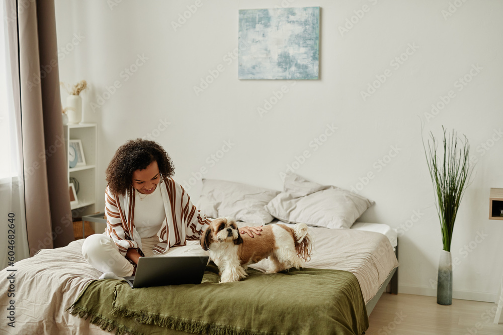 Cozy portrait of black young woman relaxing at home sitting on bed with cute pet dog and using laptop, copy space Cozy portrait of black young woman relaxing at home sitting on bed with cute pet dog and using laptop, copy space