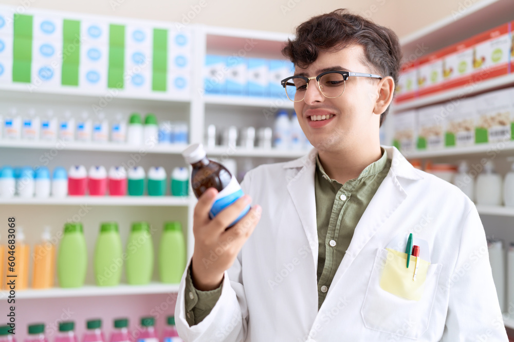Non binary man pharmacist smiling confident holding medication bottle at pharmacy Non binary man pharmacist smiling confident holding medication bottle at pharmacy