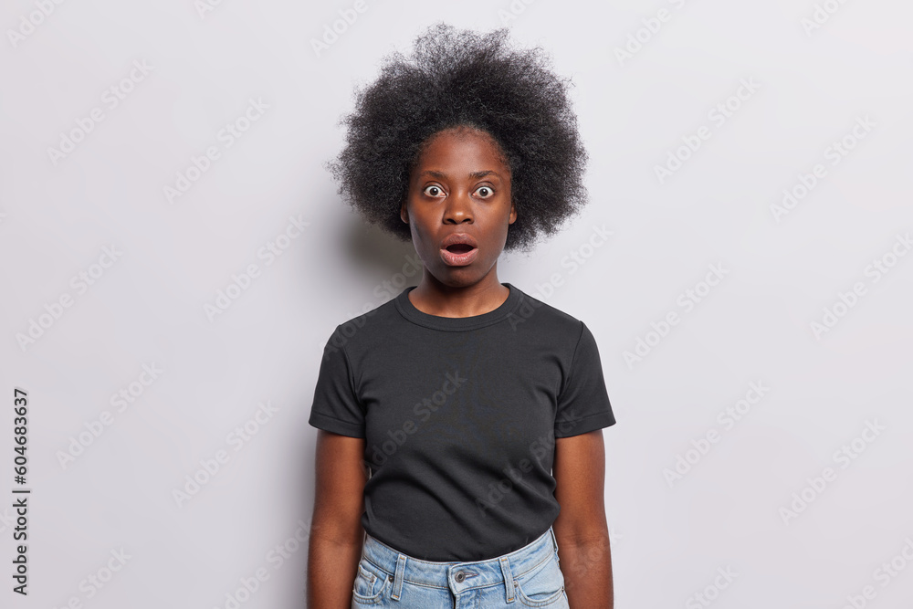 Frightened Afro woman with dark bushy hair whitnesses crime stares with shocked scared expression keeps mouth opened dressed in black t shirt jeans isolated over white background. No it cant be true - obrazy, fototapety, plakaty 