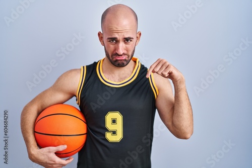 Young bald man with beard wearing basketball uniform holding ball pointing down looking sad and upset, indicating direction with fingers, unhappy and depressed.