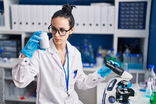 Young caucasian woman scientist using microscope holding pills bottle at laboratory