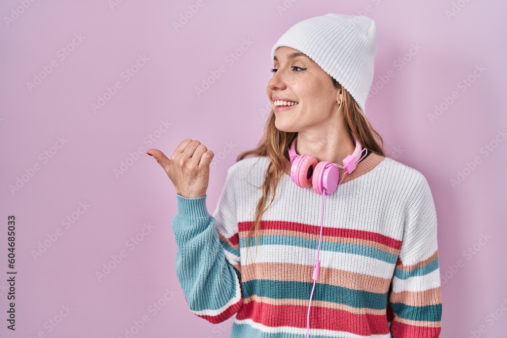 Young blonde woman standing over pink background smiling with happy face looking and pointing to the side with thumb up. Young blonde woman standing over pink background smiling with happy face looking and pointing to the side with thumb up.