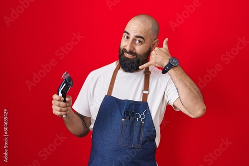 Young hispanic man with beard and tattoos wearing barber apron holding razor smiling doing phone gesture with hand and fingers like talking on the telephone. communicating concepts. © Krakenimages.com