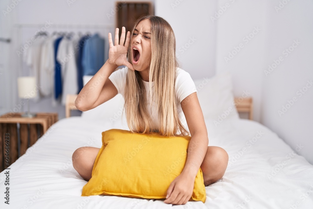 Young blonde woman sitting on the bed with pillow at home shouting and screaming loud to side with hand on mouth. communication concept. Young blonde woman sitting on the bed with pillow at home shouting and screaming loud to side with hand on mouth. communication concept.