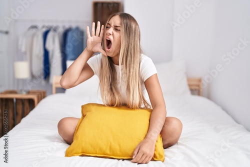 Young blonde woman sitting on the bed with pillow at home shouting and screaming loud to side with hand on mouth. communication concept.