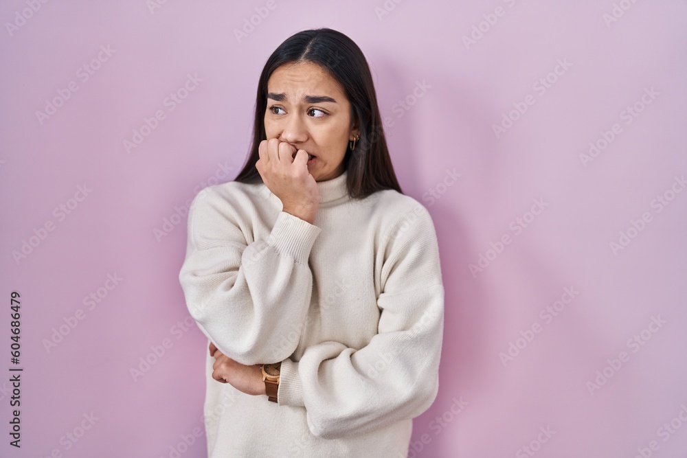 Young south asian woman standing over pink background looking stressed and nervous with hands on mouth biting nails. anxiety problem.