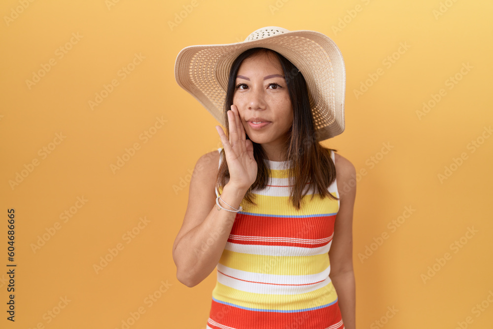 Middle age chinese woman wearing summer hat over yellow background hand on mouth telling secret rumor, whispering malicious talk conversation Middle age chinese woman wearing summer hat over yellow background hand on mouth telling secret rumor, whispering malicious talk conversation